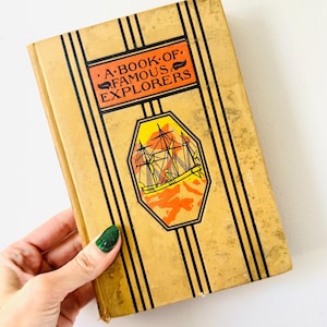 May include: A vintage book titled "A Book of Famous Explorers" with a tan cover and black vertical stripes. The cover features a ship illustration in a yellow and orange octagon. The book is held by a hand with a green glitter nail polish.