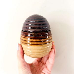 May include: A handmade ceramic jar with a lid. The jar is egg-shaped and features horizontal bands of color, transitioning from a light tan base to shades of brown and dark brown at the top. The lid mirrors the top portion of the jar.