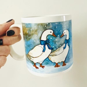 May include: White ceramic mug featuring two geese with blue bows, set against a wintery blue and white background. The geese are depicted in a whimsical, illustrated style. The mug has a white handle.