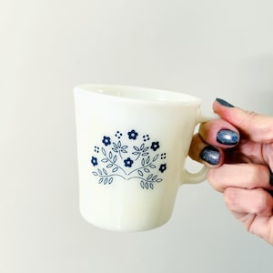 May include: Cream-colored mug with a blue floral design. The mug features a curved handle and a slightly angled rim. The floral pattern includes small flowers and leaves, adding a touch of vintage charm to this kitchenware item.