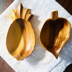 May include: Two wooden serving bowls. One is shaped like a pineapple, the other like a leaf. Both are made of light brown wood with visible grain patterns. The bowls are empty and sit on a white, patterned cloth.