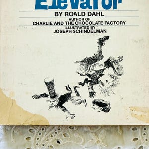 Puede incluir: Un libro de tapa dura vintage titulado Charlie and the Great Glass Elevator de Roald Dahl. La cubierta de color blanco hueso presenta ilustraciones en tinta negra de dos figuras en movimiento, con el autor Roald Dahl y el ilustrador Joseph Schindelman.