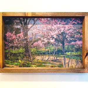 May include: An oil painting of a landscape featuring trees with pink blossoms, set within a rustic wooden frame. The artwork depicts a serene scene with a pathway and a body of water, evoking a sense of tranquility and natural beauty.