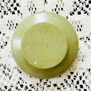 May include: A light green, round, stone-like object with a smaller, raised circular section in the center. The object has a textured surface and appears to be handmade. The background is a white, open-weave fabric.