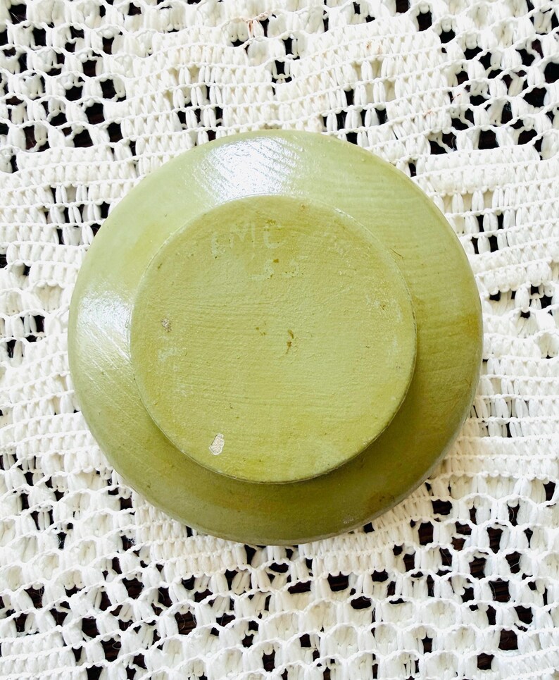 May include: A light green, round, stone-like object with a smaller, raised circular section in the center. The object has a textured surface and appears to be handmade. The background is a white, open-weave fabric.