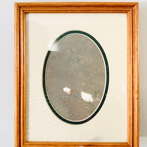 May include: A rectangular wooden picture frame with an oval opening. The frame is a light brown color, and the oval opening is surrounded by a green border. The frame is held up against a white wall.