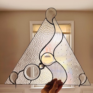 May include: A stained glass triangle with a black border. The design features clear, textured, and iridescent glass sections. The piece has a circular element and a rounded top, creating an abstract, artistic look.