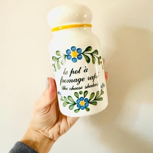 May include: White ceramic cheese shaker with a rounded top and yellow trim. The shaker is decorated with blue floral designs and green leaves. Text on the shaker reads "le pot à fromage rapé (the cheese shaker)".