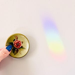May include: A small, round, vintage trinket box with a floral design. The box features a red rose with green leaves on a light green background. The box is held by a hand with blue nail polish.