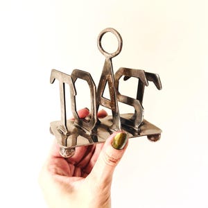 May include: Silver-toned metal toast rack with the word "TOAST" in large capital letters. The rack has a rectangular base with small ball feet and a handle at the top. A vintage kitchenware item.