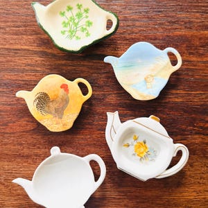 May include: A collection of five ceramic teapot-shaped tea bag holders. The holders feature various designs, including a rooster, a beach scene, a shamrock, and a floral pattern. Each holder has a handle and a spout.