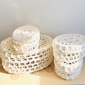 May include: A collection of off-white, crocheted round boxes in varying sizes. The boxes are stacked, showcasing the open, lacy weave of the handmade items. The boxes are likely used for storage or decoration, adding a touch of handmade charm.