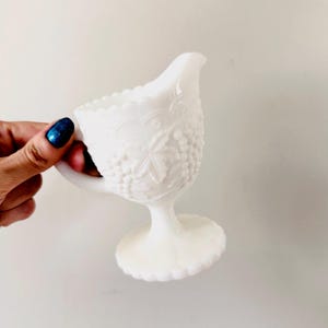 May include: A white, vintage creamer with a detailed grape and leaf pattern. The creamer has a scalloped rim, a handle, and a pedestal base. The creamer is made of opaque glass and is approximately 5 inches tall.