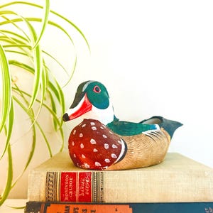 May include: A colorful ceramic duck figurine with red, white, green, and brown details. The duck is perched atop a stack of vintage books with titles like "Robinson Crusoe" and "The Postman Always Rings Twice".
