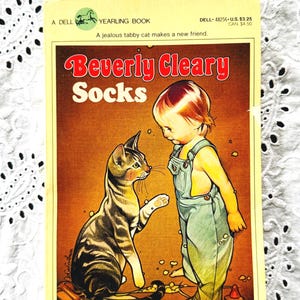 May include: A paperback book titled Socks by Beverly Cleary, published by Dell Yearling. The cover features a brown tabby cat facing a young child wearing green overalls. The text reads A Dell Yearling Book, A jealous tabby cat makes a new friend.