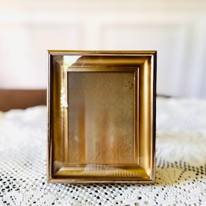 May include: A rectangular picture frame with a metallic gold finish and a beveled border. The frame features a clear glass front panel and a smooth, reflective surface that catches light, creating a bright sheen along the edges and corners.