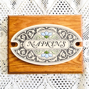 May include: A wooden plaque with an oval ceramic insert. The insert features the word "NAPKINS" in a decorative font, surrounded by a floral and lace design in black, white, and blue. The plaque is made of light brown wood.