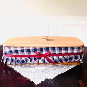 May include: A rectangular wicker basket with a wooden lid. The basket is lined with blue and white plaid fabric, trimmed with a red ribbon and a matching bow. The basket is sitting on a white lace doily.