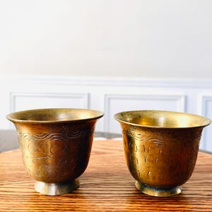May include: Two antique brass cups with etched floral designs. The cups have a flared rim and a small, round base. The brass has a warm, golden hue, and the cups are displayed on a wooden surface.