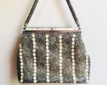 May include: A silver beaded handbag featuring vertical rows of small white floral-shaped beads. The bag has a metallic silver beaded handle and a rectangular frame top accented with iridescent white rectangular panels along the closure.