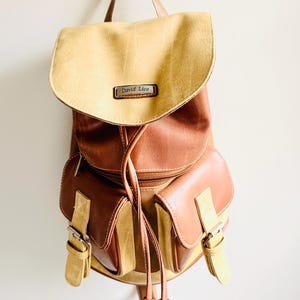 May include: A two-tone leather backpack with a flap closure and two front pockets. The top flap is tan, and the body is brown. The backpack has adjustable straps and a drawstring closure. The brand name "David Lico" is visible on the front.