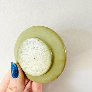 May include: A light green, circular object with a smaller, lighter green circle on top. The smaller circle has the letters "IMC" and the number "55" etched into it. The object is held by a hand with blue nail polish.