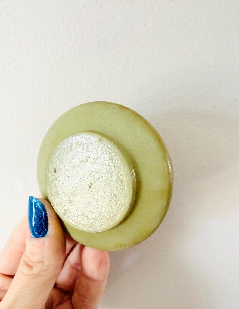 May include: A light green, circular object with a smaller, lighter green circle on top. The smaller circle has the letters "IMC" and the number "55" etched into it. The object is held by a hand with blue nail polish.