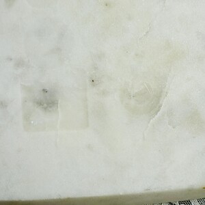 May include: Close-up of a white marble surface with subtle gray veining and a slightly textured appearance. The marble has a natural, polished look, with a hint of translucence. The bottom edge of the marble is visible, and a lace fabric is at the bottom.