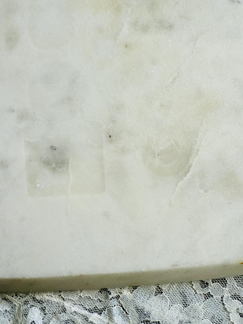 May include: Close-up of a white marble surface with subtle gray veining and a slightly textured appearance. The marble has a natural, polished look, with a hint of translucence. The bottom edge of the marble is visible, and a lace fabric is at the bottom.