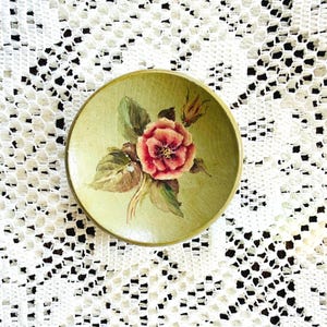 May include: A small, round, hand-painted wooden dish with a light green background. The dish features a detailed painting of a red and pink rose with green leaves. The dish is likely used for decorative purposes.