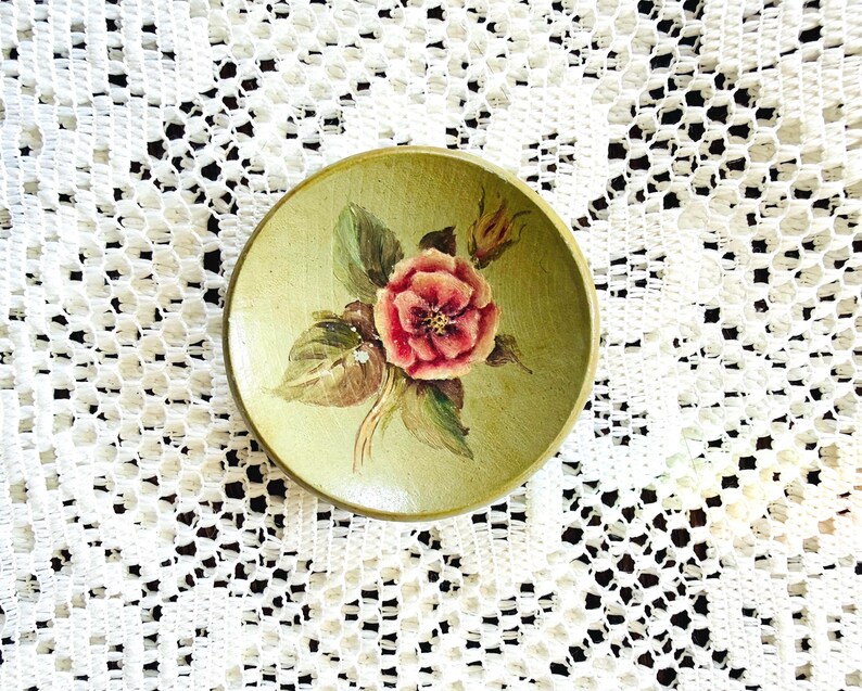 May include: A small, round, hand-painted wooden dish with a light green background. The dish features a detailed painting of a red and pink rose with green leaves. The dish is likely used for decorative purposes.