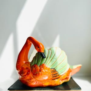 May include: A ceramic swan figurine with orange, green, and black accents. The swan has a long, curved neck and is perched on a black base. The wings are green with detailed feather patterns.