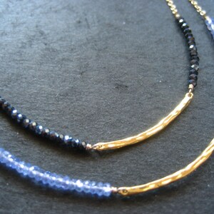 以下が含まれることがあります： 青と黒の宝石ビーズが付いた2本のゴールドネックレス。ネックレスは重ねて着用し、テクスチャ加工されたゴールドバーペンダントが付いています。