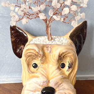 May include: A ceramic planter shaped like a dog head with tan and brown fur, featuring a wire tree sculpture with small clear crystal leaves emerging from the top. The base of the tree is surrounded by small white stones inside the planter opening.