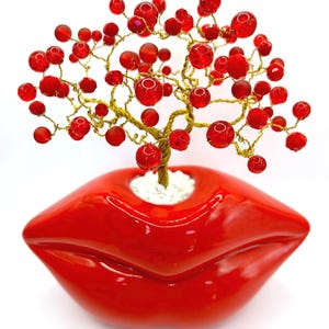 May include: A decorative tree sculpture with a gold wire trunk and branches, adorned with numerous red beads. The tree is planted in a glossy red lip-shaped planter, creating a unique and artistic display.
