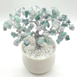 Puede incluir: Una escultura de árbol decorativa con tronco y ramas de alambre, adornada con piedras preciosas verdes, blancas y rosas. El árbol está plantado en una pequeña maceta de cerámica blanca llena de guijarros blancos.