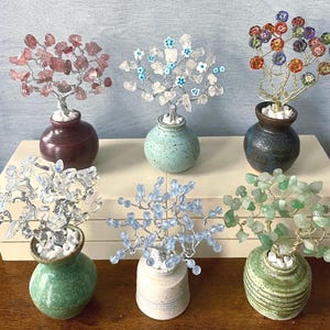 May include: Seven small potted trees made of wire and various colored gemstones. The trees are in different colored ceramic pots. The trees are arranged on a white surface.
