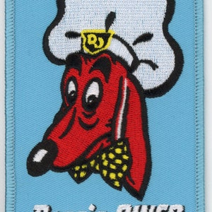 May include: A blue and white embroidered patch with a red cartoon dog wearing a white chef's hat. The dog has a black nose and eyes. The text "Doggie Diner" is embroidered in white below the dog.