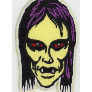 May include: Embroidered patch of a green-skinned woman with purple hair and red eyes. The woman has a sinister expression and appears to be a monster.