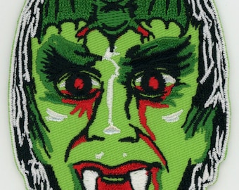 Puede incluir: Parche bordado verde de la cara de un monstruo con colmillos y sangre goteando. Un murciélago verde con ojos rojos está posado en la frente del monstruo.
