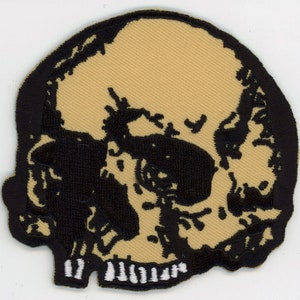 May include: A black and tan embroidered patch of a skull with a white outline around the bottom of the jaw.