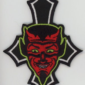 May include: A red devil face with green eyes and a black outline is embroidered on a black cross with white trim. The cross is outlined in green.