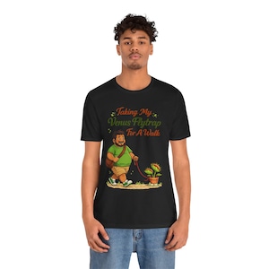 May include: Black t-shirt with a cartoon illustration of a person walking a Venus flytrap on a leash. The text "Taking My Venus Flytrap For A Walk" is above the illustration. The person is wearing a green shirt and khaki shorts.
