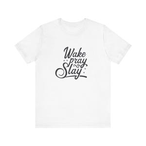 Op de afbeelding: Wit t-shirt met de tekst "Wake Pray Slay" in zwart script. De tekst staat in het midden van de voorkant van het shirt. Het shirt heeft korte mouwen en een ronde hals.