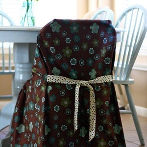 May include: A chair cover with a brown floral pattern, featuring teal and green flowers. The cover is tied with a cream-colored ribbon with black polka dots. The chair is part of a dining set.