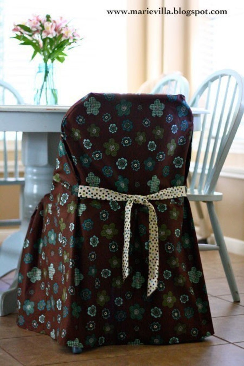 May include: A chair cover with a brown floral pattern, featuring teal and green flowers. The cover is tied with a cream-colored ribbon with black polka dots. The chair is part of a dining set.
