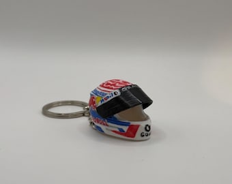 May include: A miniature racing helmet keychain featuring a white, red, and blue color scheme with various sponsor logos and text including Mobil 1 and Oracle. The helmet has a black visor and is attached to a circular silver metal keyring.