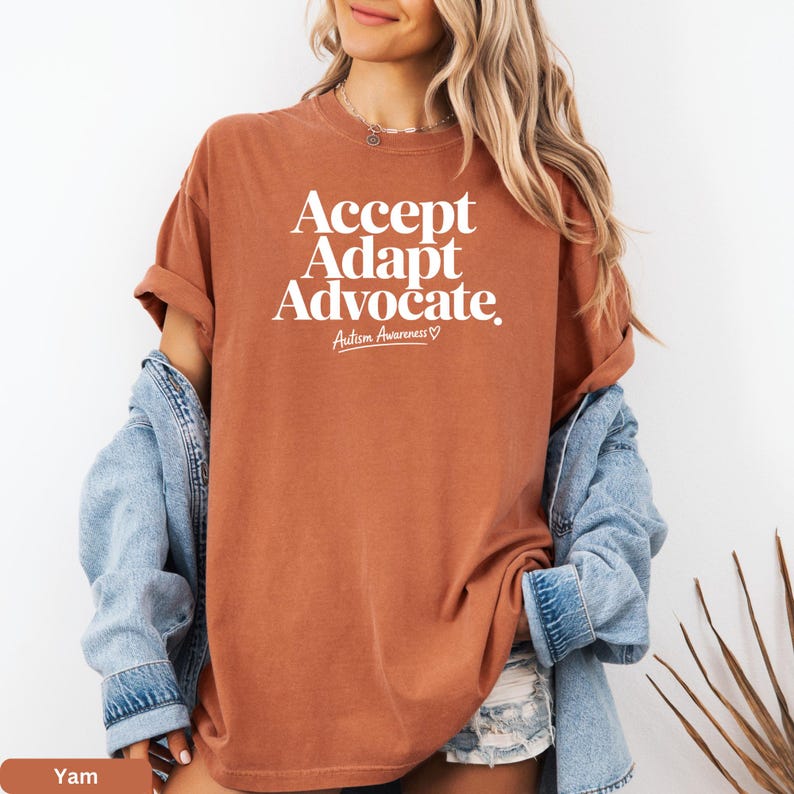 A woman wearing a brown t-shirt with the text "Accept Adapt Advocate" printed on it, standing against a white background.