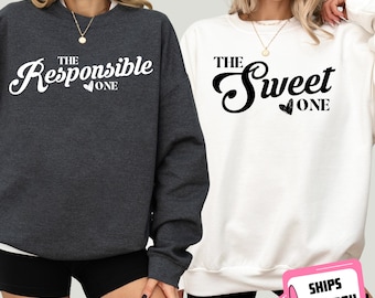 May include: Two sweatshirts with a matching design. One sweatshirt is dark gray with the text "The Responsible One" in white. The other sweatshirt is white with the text "The Sweet One" in black.