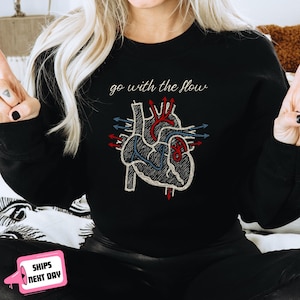 May include: Black sweatshirt featuring an embroidered anatomical heart design with red and blue accents. The phrase "go with the flow" is written above the heart in cursive. The sweatshirt is a casual style.